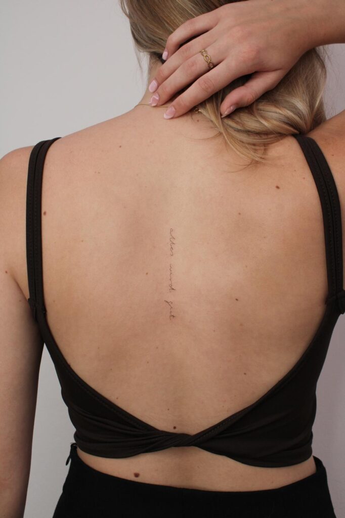 a young woman from behind, holding back her hair, showing a minimalist calligraphy tattoo on her spine